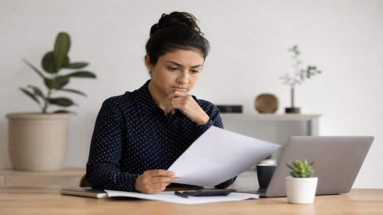 Mulher concentrada analisando documentos financeiros em uma mesa de madeira em ambiente minimalista e iluminado suavemente. Ela segura uma folha de papel enquanto observa atentamente as informações, com notebook aberto ao lado, celular, calculadora e pequenos objetos decorativos ao fundo.
